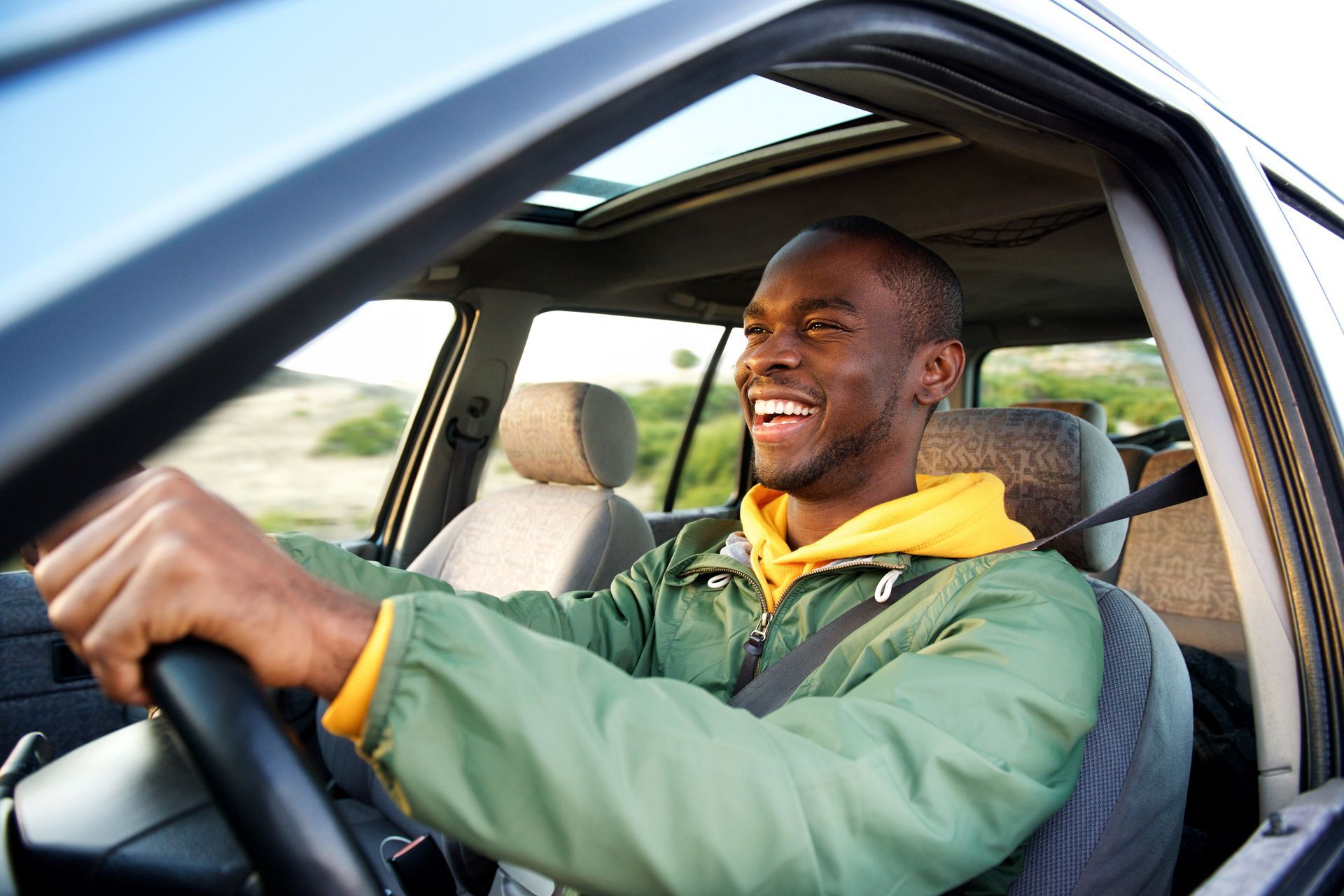 Smiling driver in green jacket and yellow hoodie driving a car with the window open