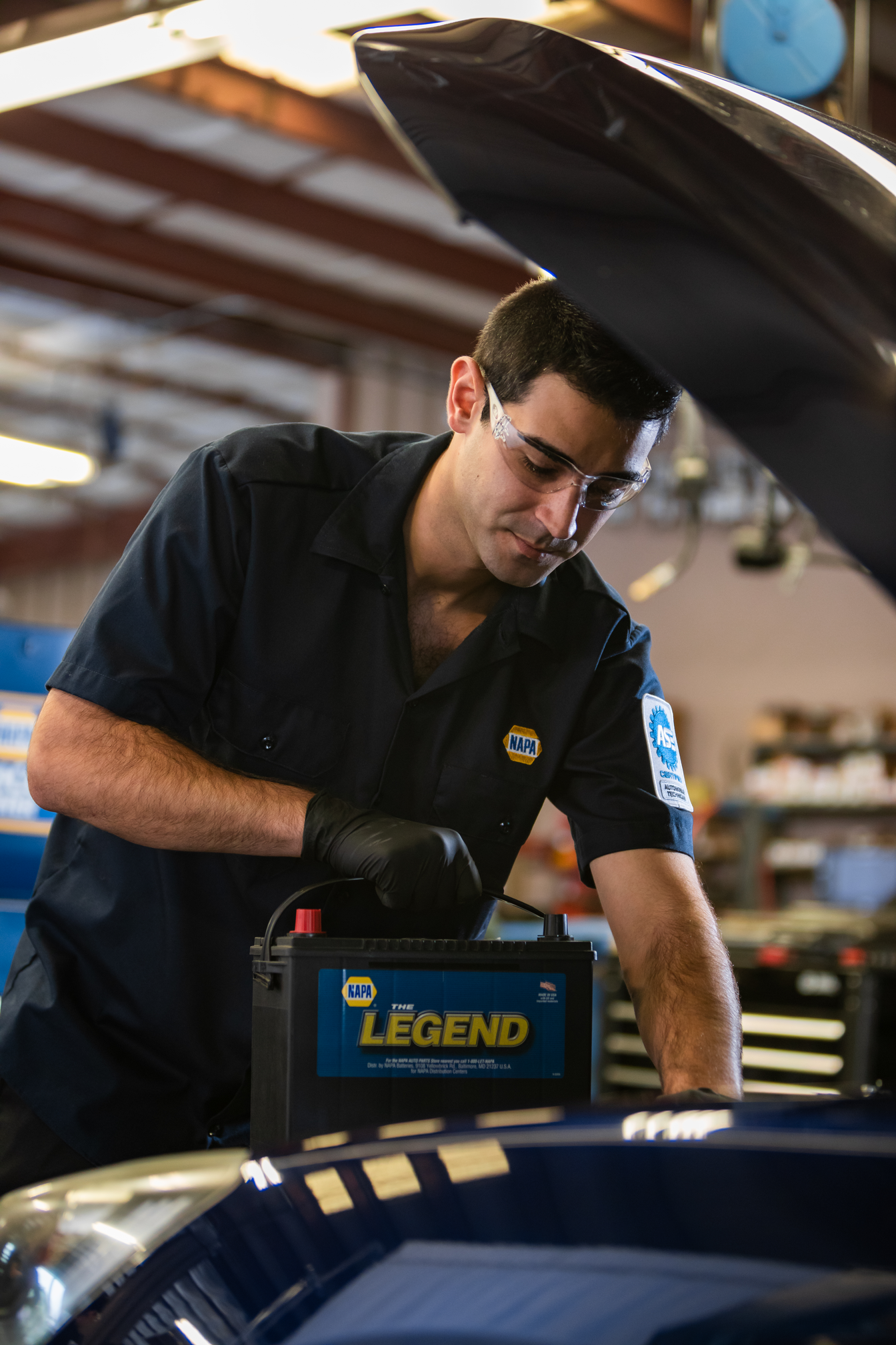 Mechanic working under a raised car hood with a battery charger in a garage