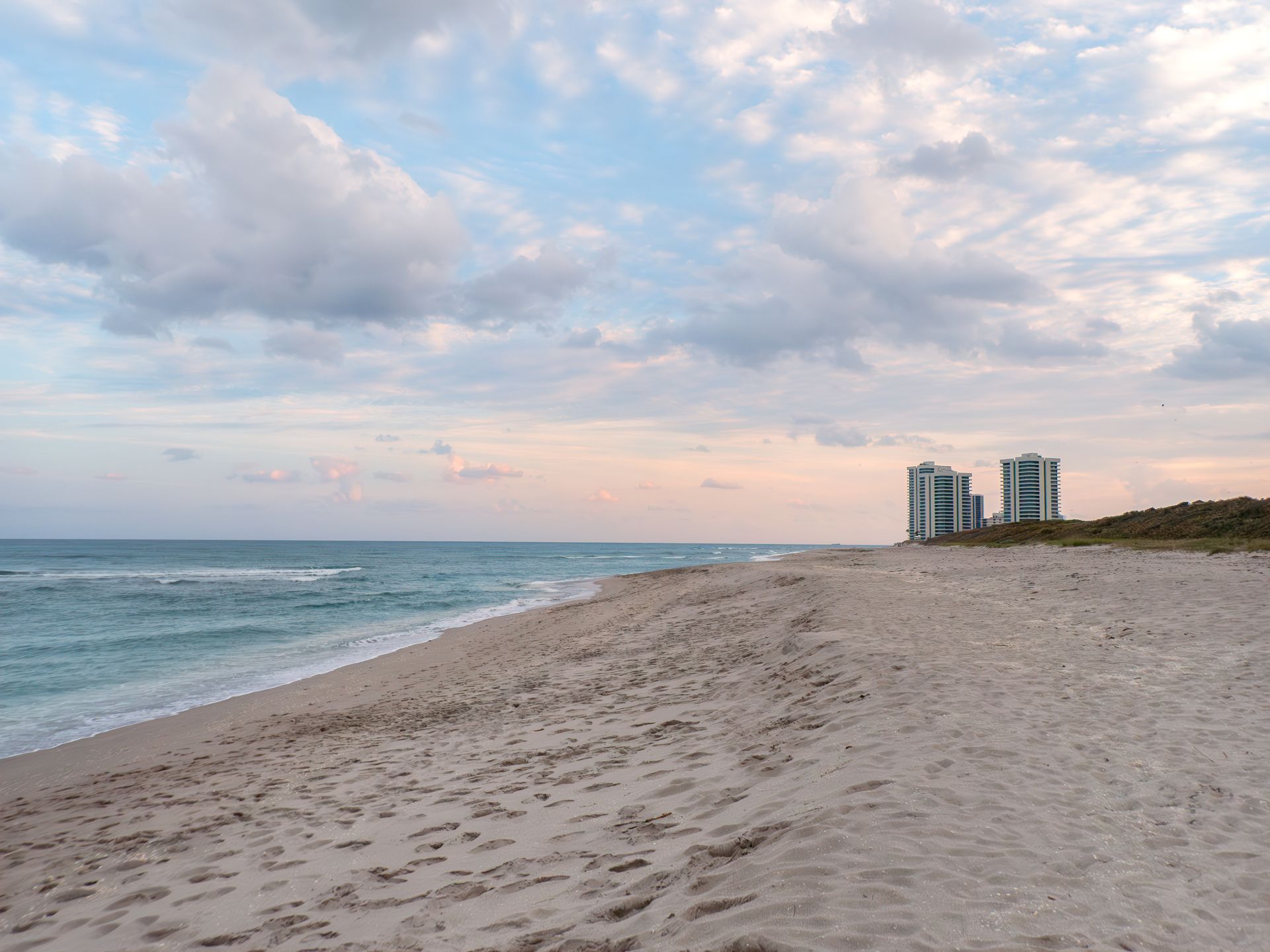 Wide sandy beach beside calm ocean under cloudy pastel sky, with distant high-rise buildings on the shore
