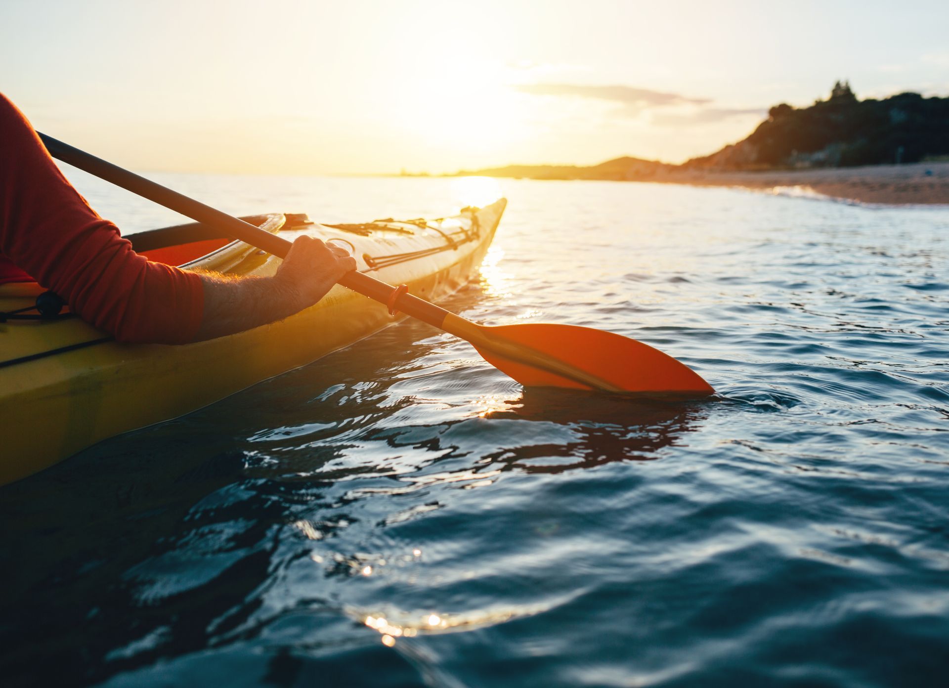 Kayak paddling on calm water at sunset, with an orange paddle and bright golden horizon