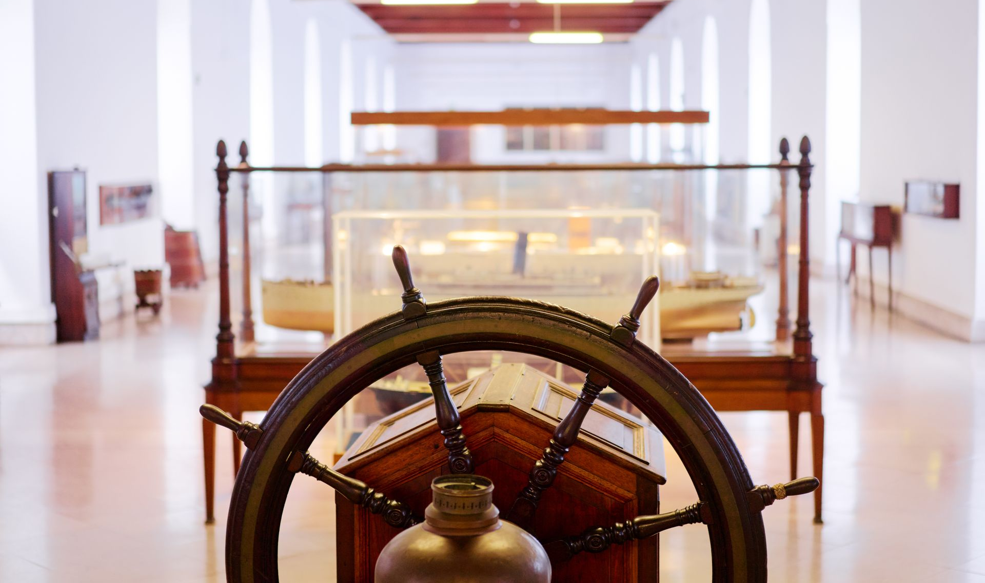 Vintage ship wheel in a bright museum gallery, with a glass display case behind it.