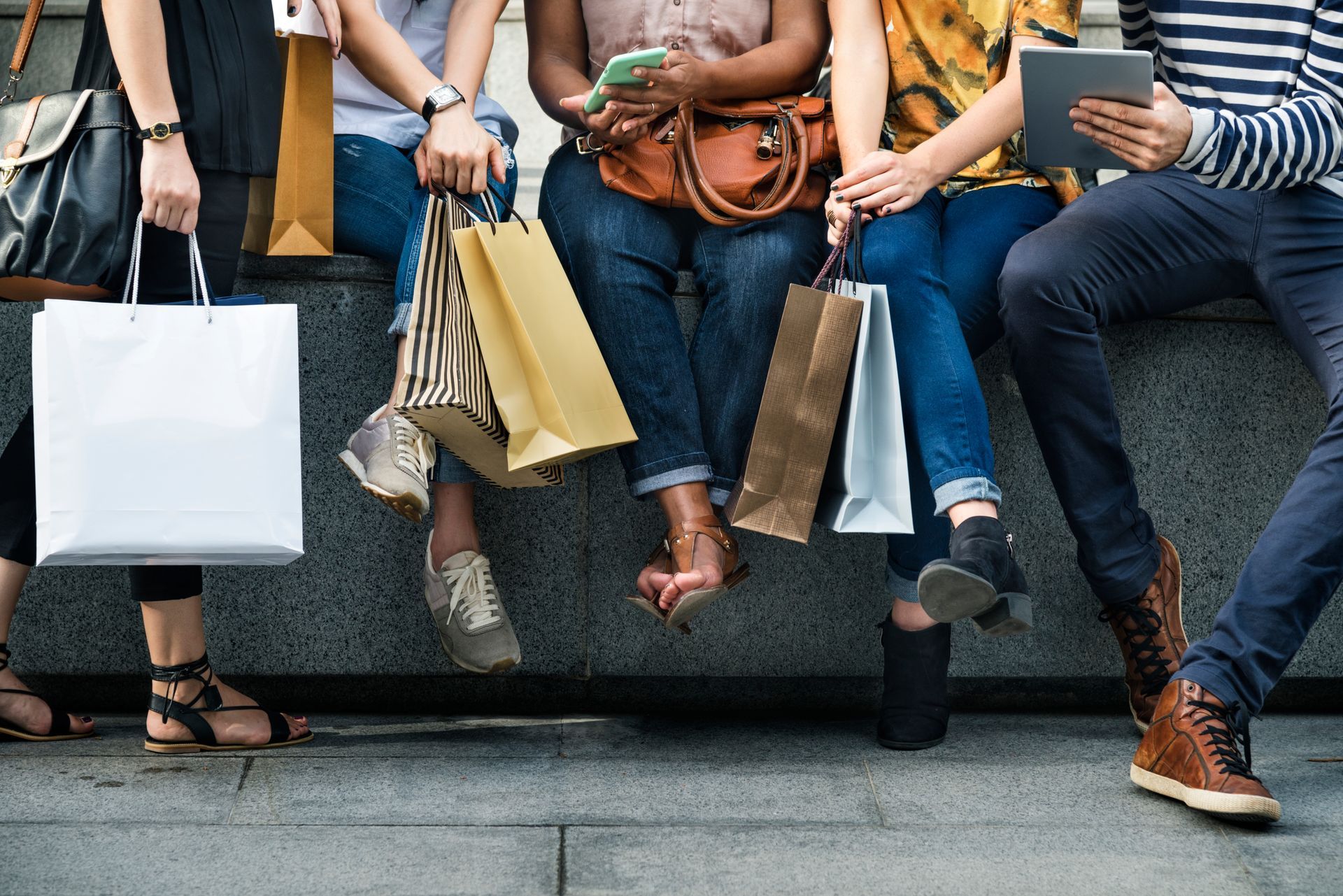 People sitting on a bench with shopping bags and takeaway cups, legs dangling over a step.