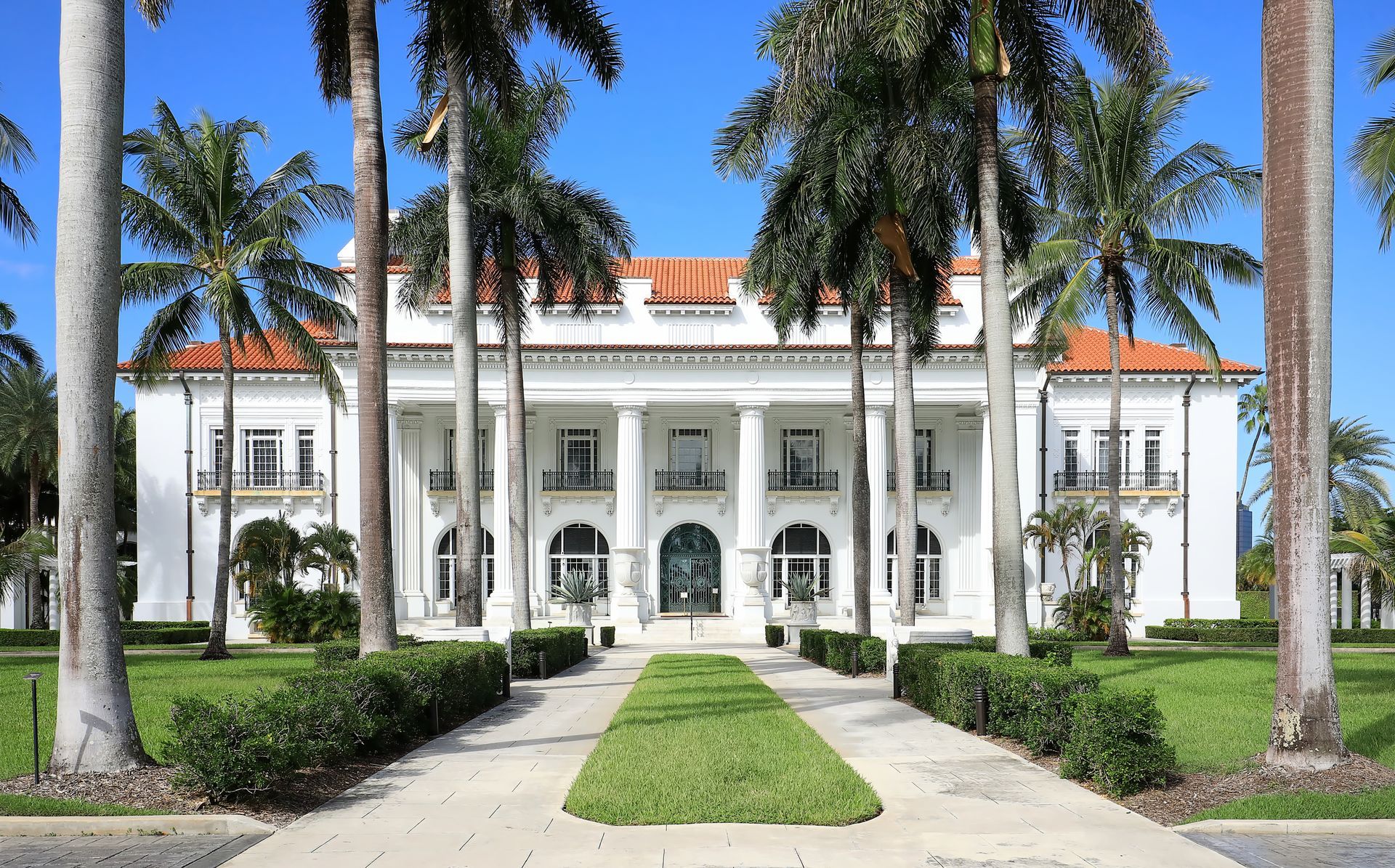 Grand white mansion with red roof, framed by tall palm trees and a central walkway