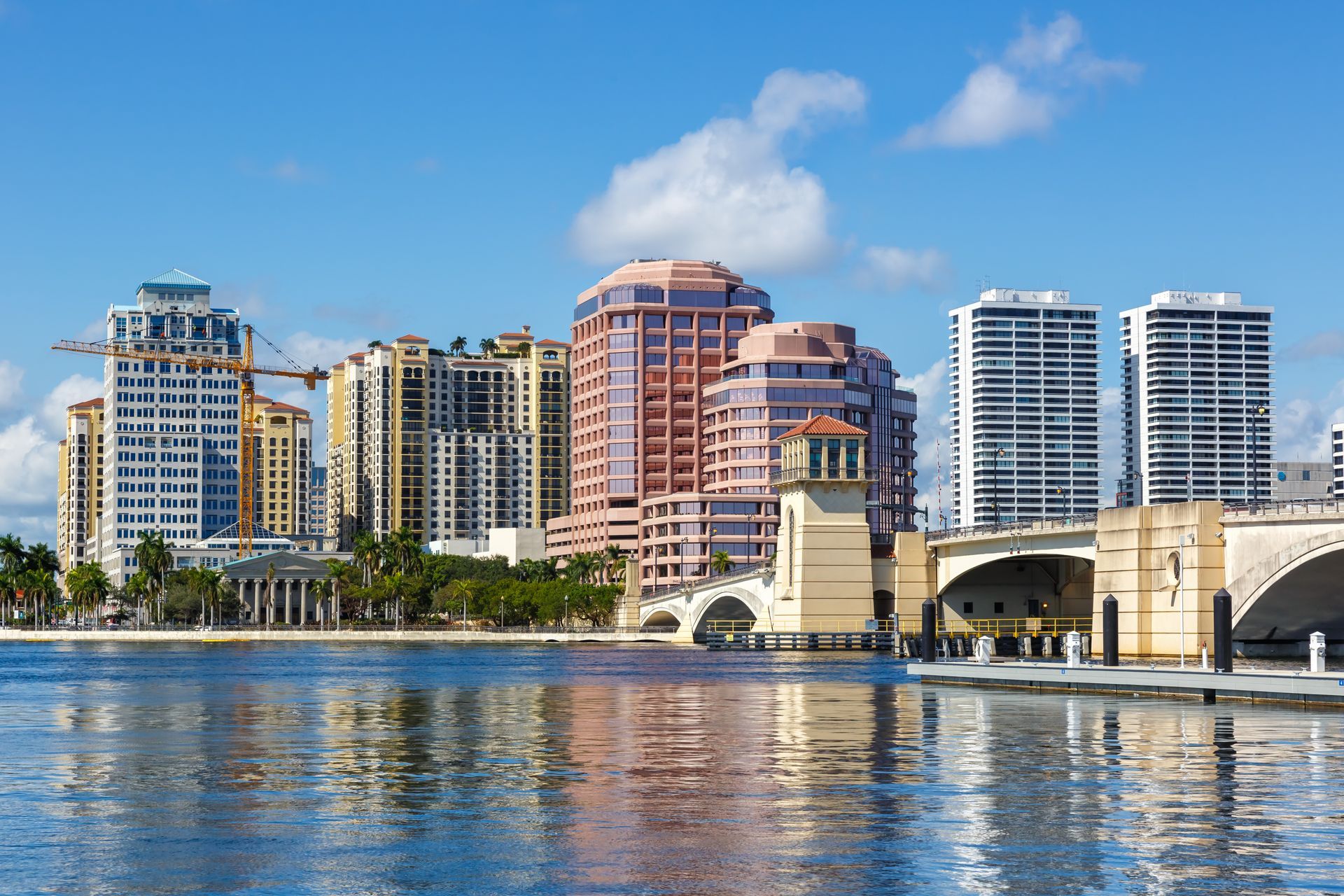 Waterfront skyline with high-rise apartments, a bridge, and reflections on calm water under a blue sky