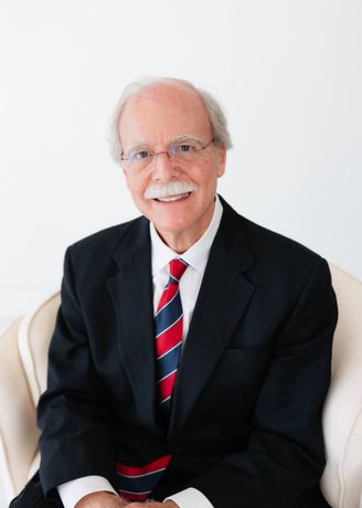 Floyd Don Davis in a black suit and patterned tie, seated on a white chair against a light background.