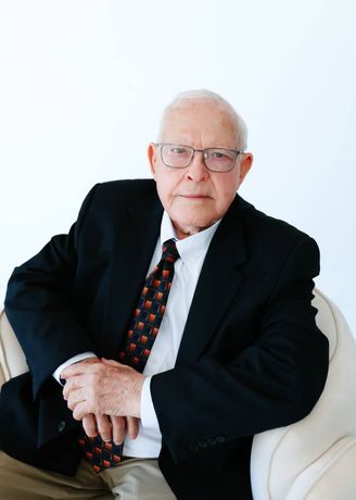 Floyd Don Davis in a black suit and patterned tie, seated on a white chair against a light background.