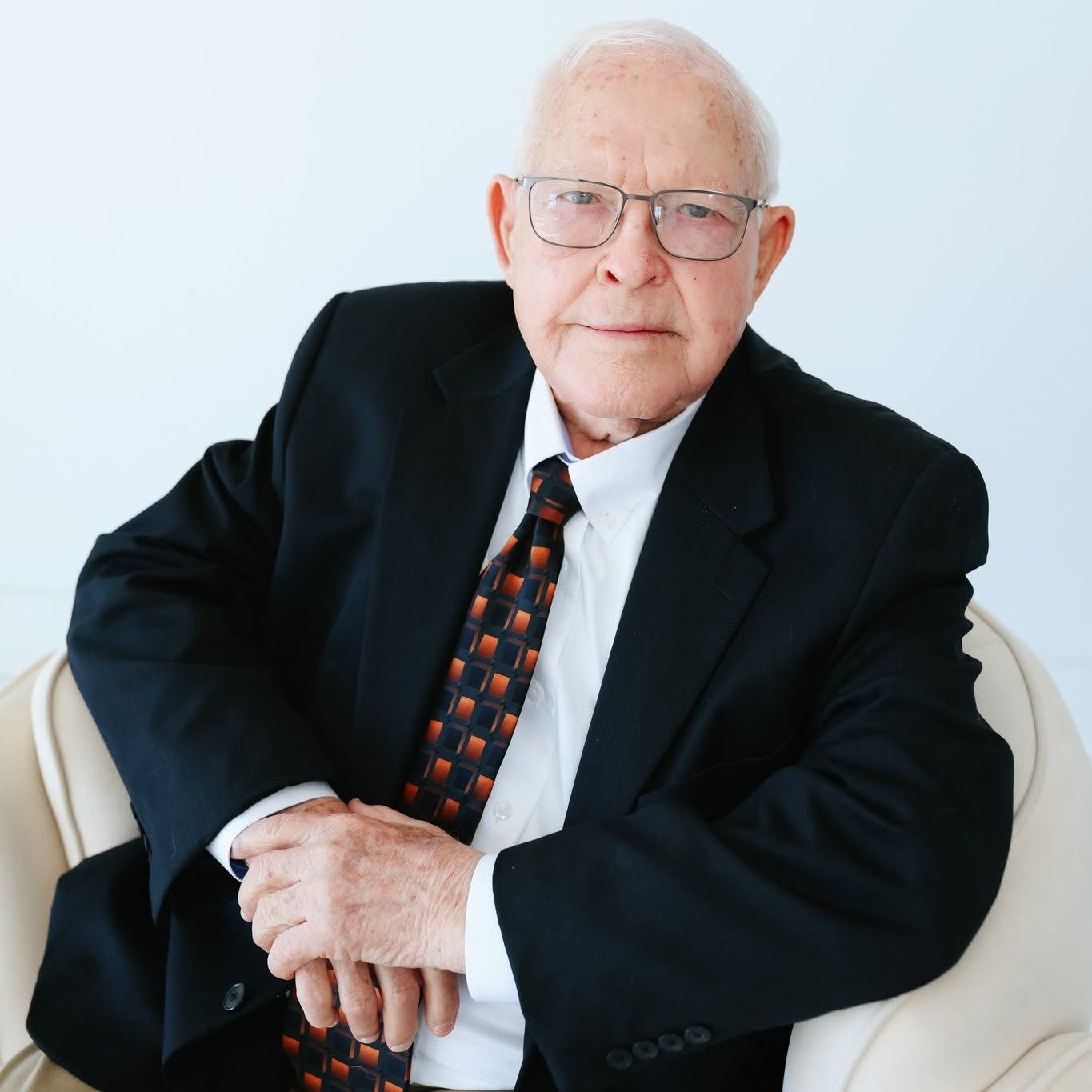 Floyd Don Davis in a black suit and patterned tie, seated on a white chair against a light background