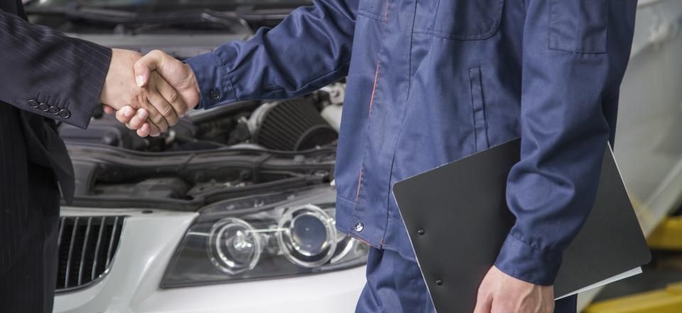 A person in a blue uniform holding a clipboard shakes hands with another person in front of an open car hood.