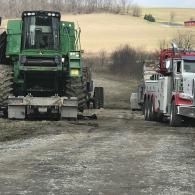 A green John Deere combine being towed by a red and white heavy-duty tow truck on a rural dirt path.