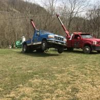 A red tow truck lifts a blue pickup truck in a grassy field in front of a wooded hillside.