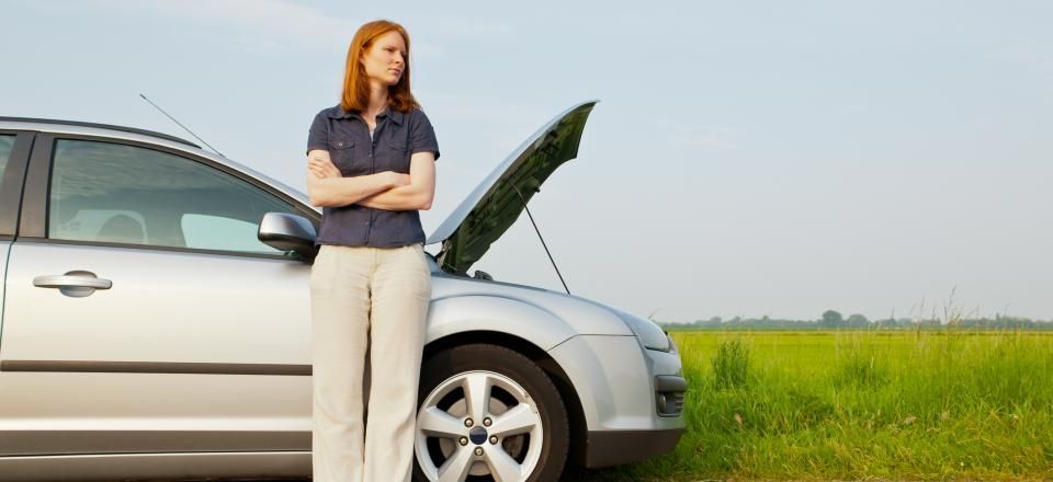 A woman with crossed arms leans against a silver car with its hood up on the side of a road near a grassy field.