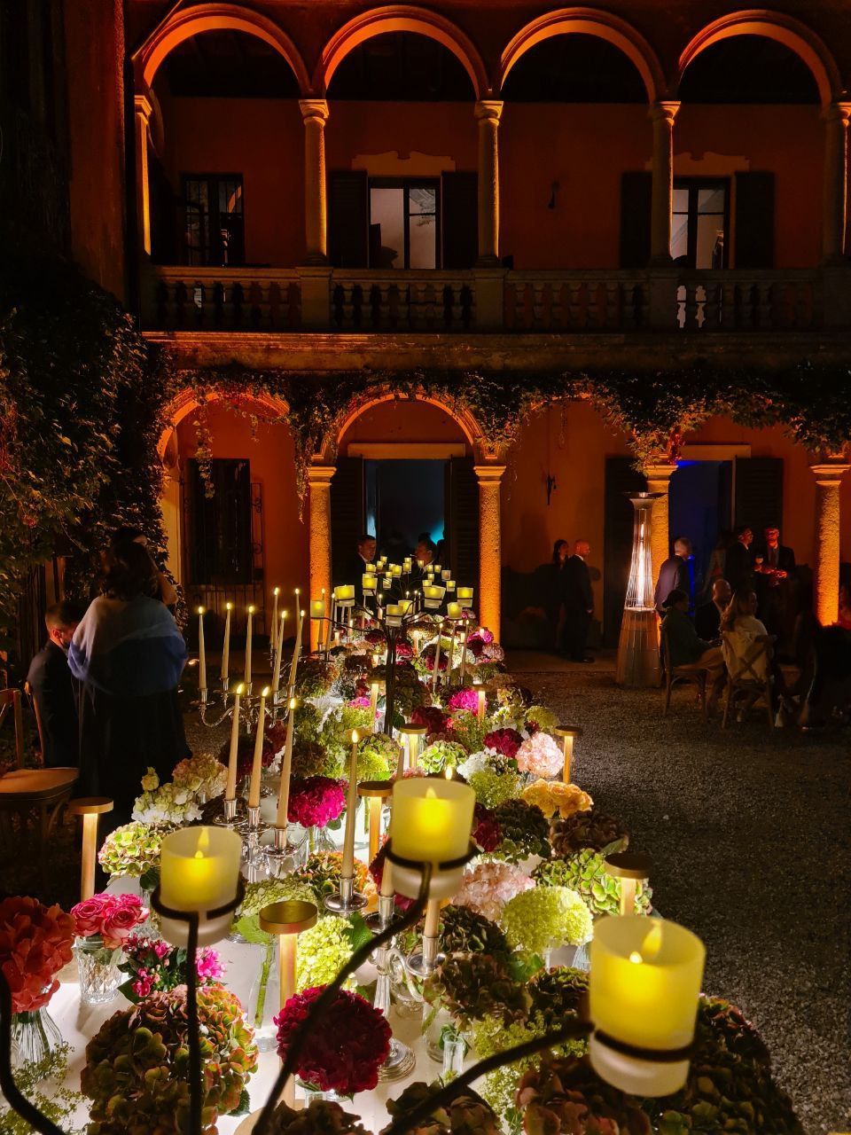 Un lungo tavolo apparecchiato per la cena, decorato con fiori e candele, sotto un arco ricoperto di vite.