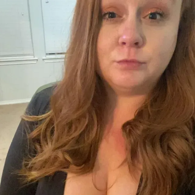 Close-up selfie of a woman with long auburn hair in a black top, indoors, with a neutral expression