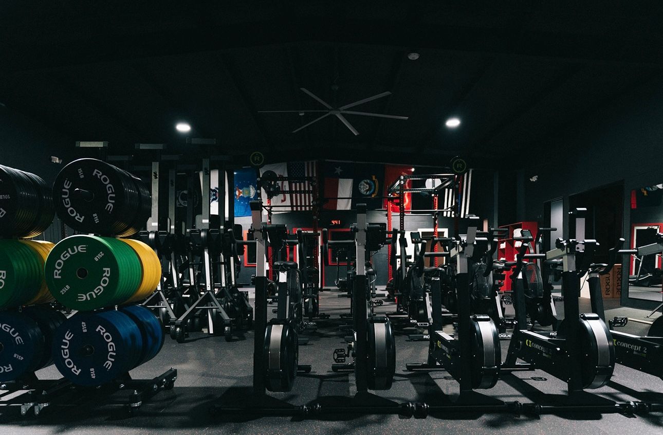 Gym interior with weight plates, rowing machines, and weightlifting equipment. Dark setting with black and red accents.