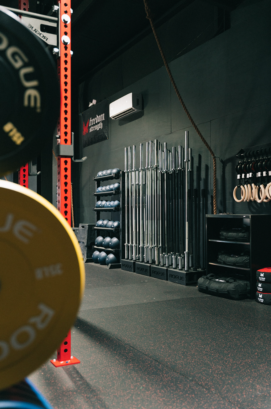 Weightlifting gym: Barbells and weights stand against a dark wall. A red power rack is in the background.