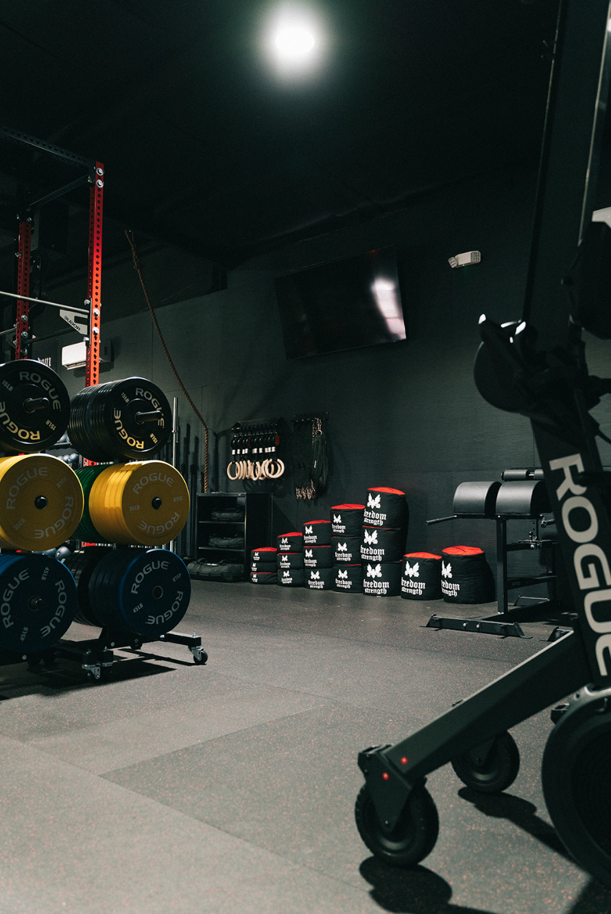 Dark gym with weight plates, barbells, plyo boxes, and a Rogue rower.