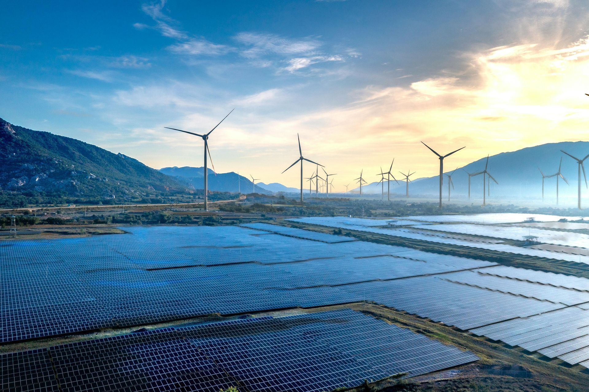Coastal wind turbines above blue reflective solar panels at sunrise, with mountains in the background