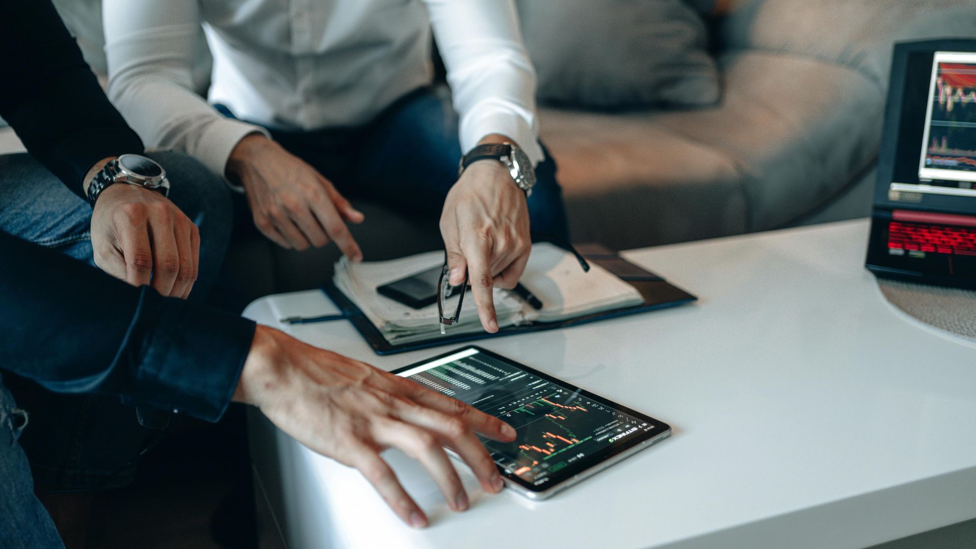 Two people review financial charts on a tablet and clipboard at a desk, with a laptop showing market data.