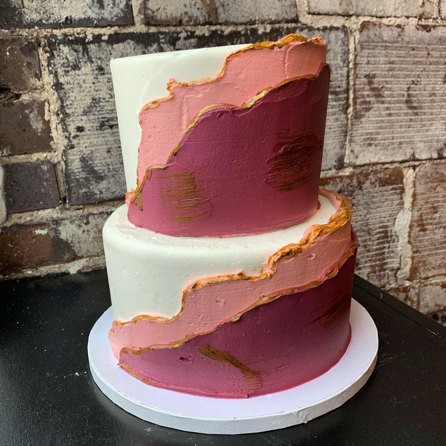 Two-tier pink and white cake with gold accents on a white stand against a brick wall