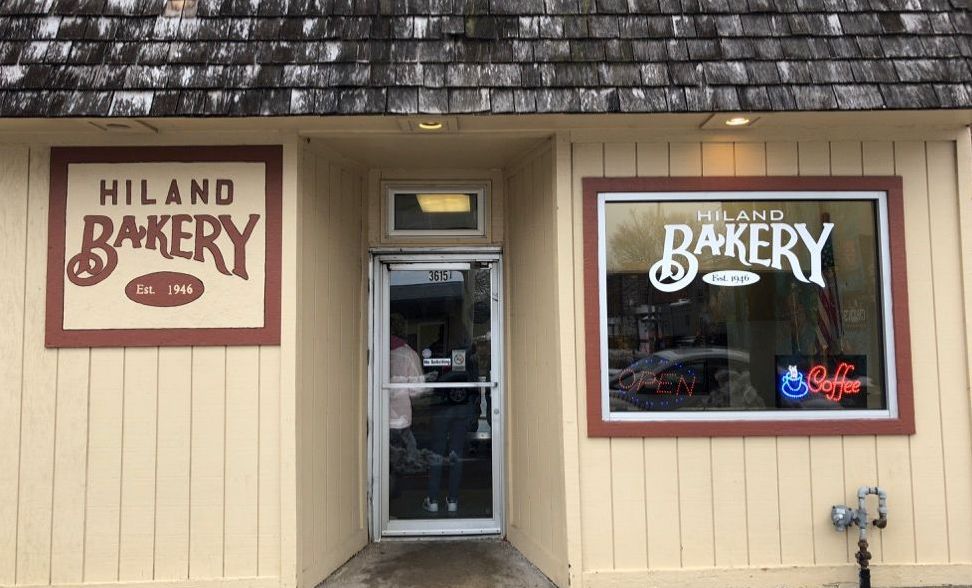 Storefront of Hiland Bakery with beige siding, brown trim, and a glass door between two signs