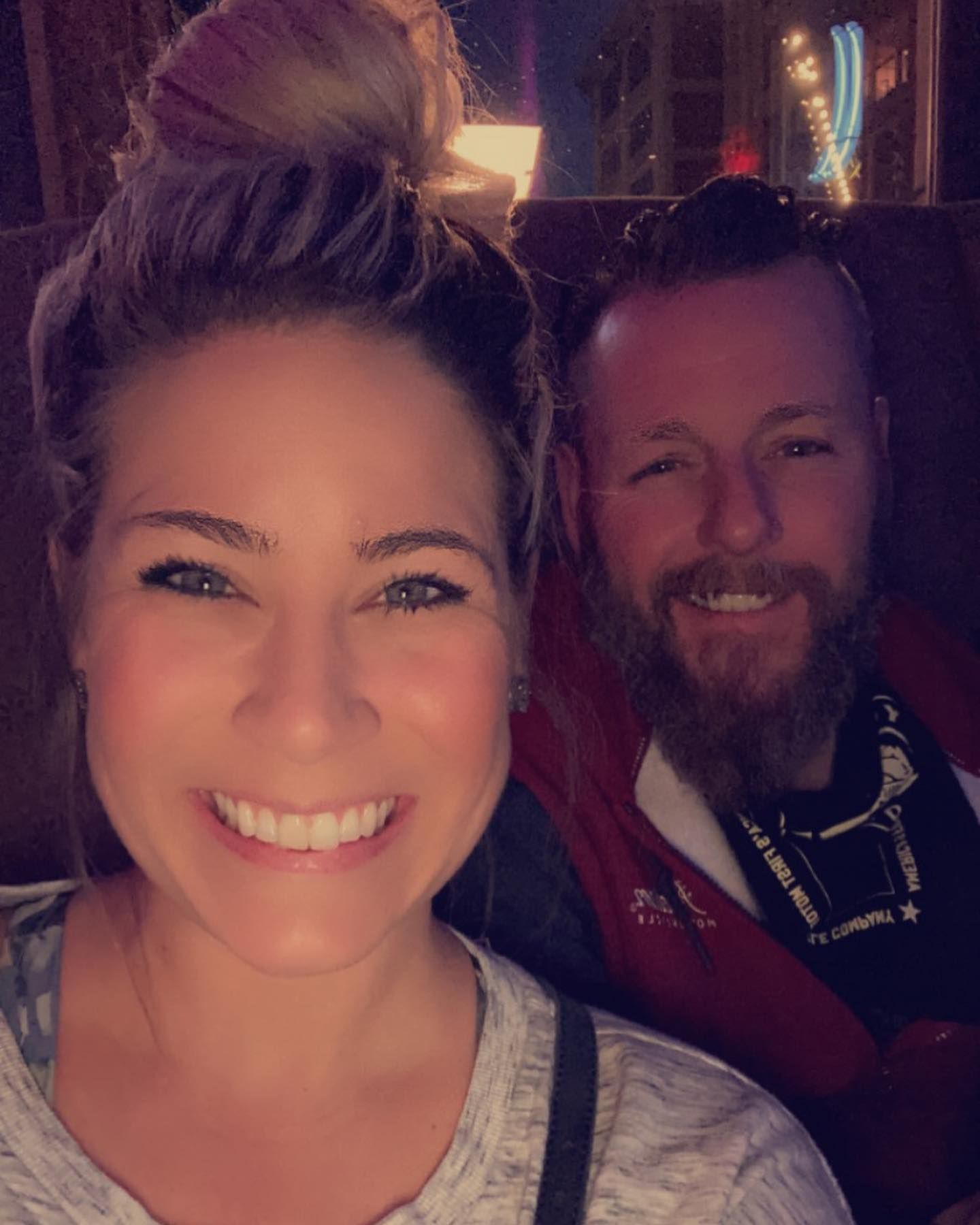 Two smiling people seated together in a dimly lit setting, one taking a selfie in front.
