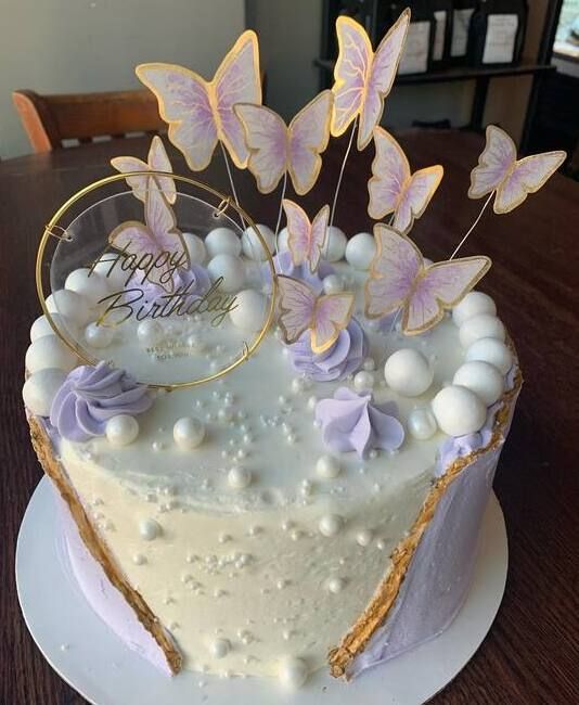 White birthday cake with purple butterflies, pearls, and a gold “Happy Birthday” topper on a wooden table
