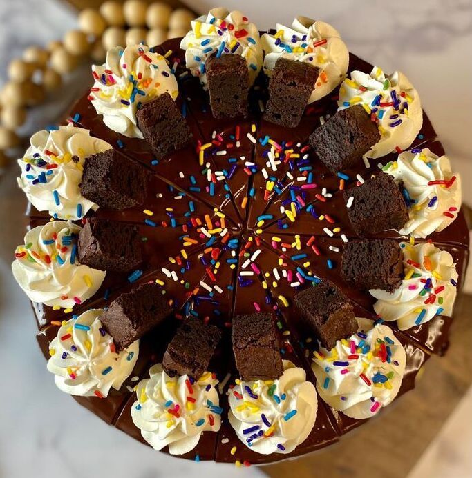 Chocolate cake topped with whipped cream, brownie squares, and colorful sprinkles on a platter