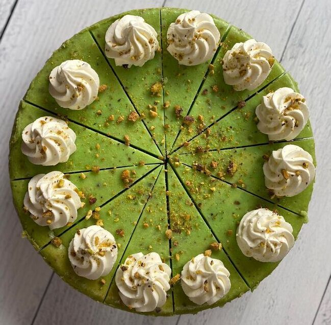 Round green cake topped with whipped cream swirls and pistachio crumbs, sliced into wedges