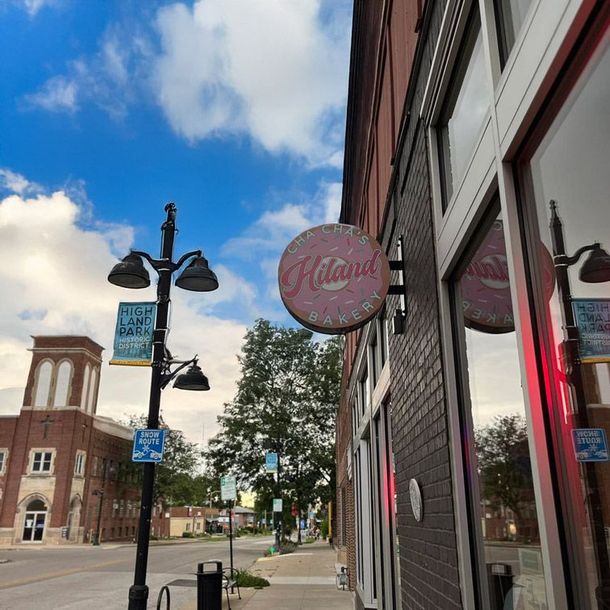 Downtown sidewalk beside storefronts, lamp posts, and a pink circular sign under a blue sky