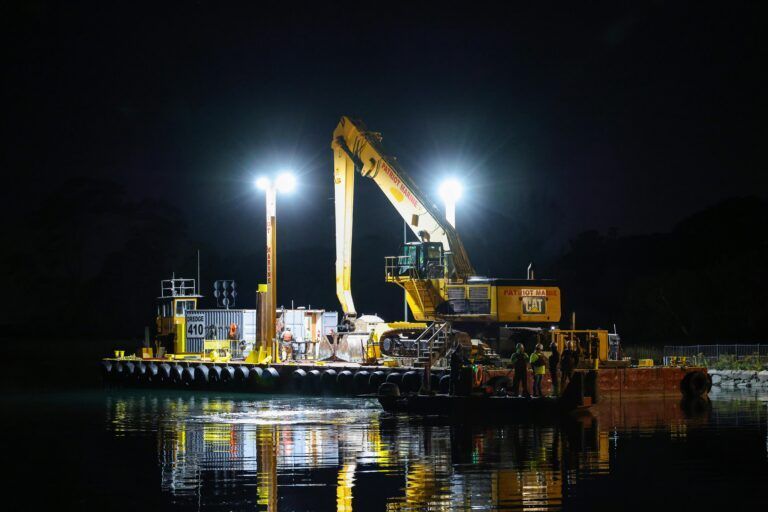 Nighttime dredging vessel lit by bright floodlights on calm water, with reflections shimmering below.