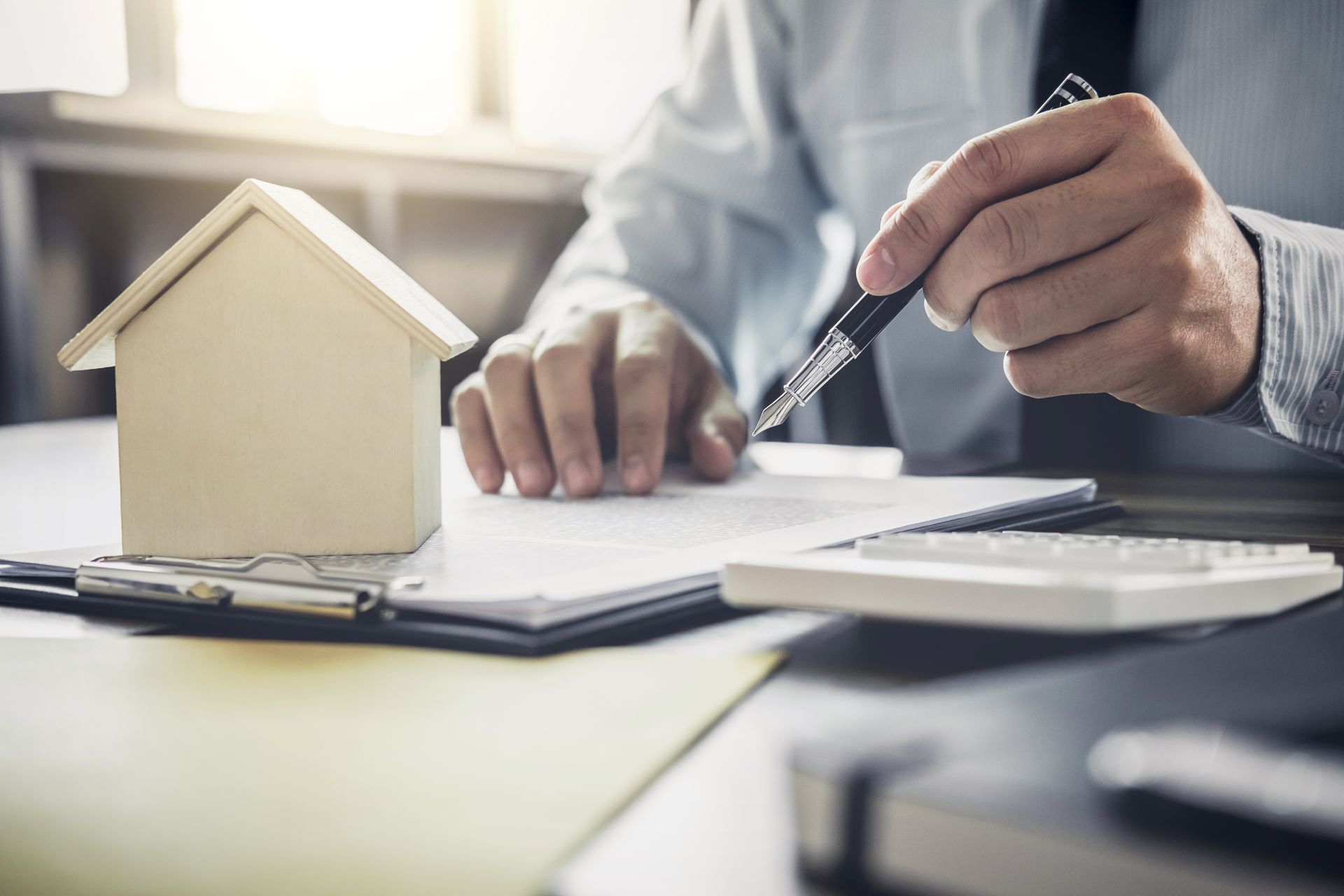Hands writing on documents beside a small house model and calculator on a desk