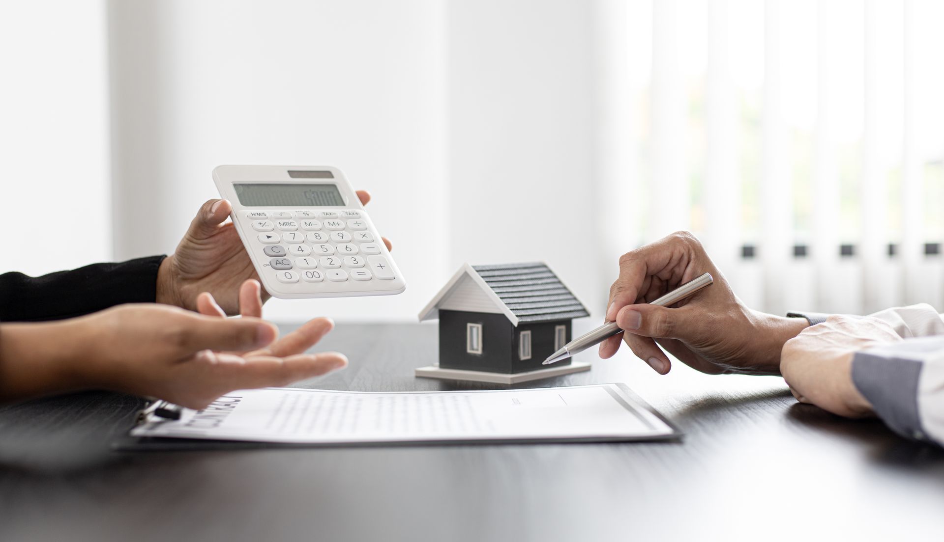 Hands reviewing a house model and calculator over paperwork at a desk, discussing finances