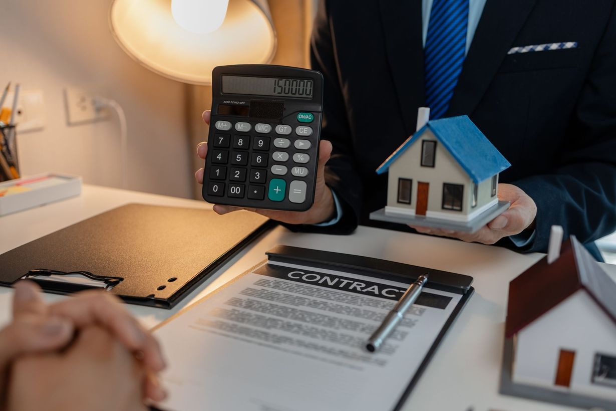 Calculator and house models on a desk with a signed finance document and a suited person in the background