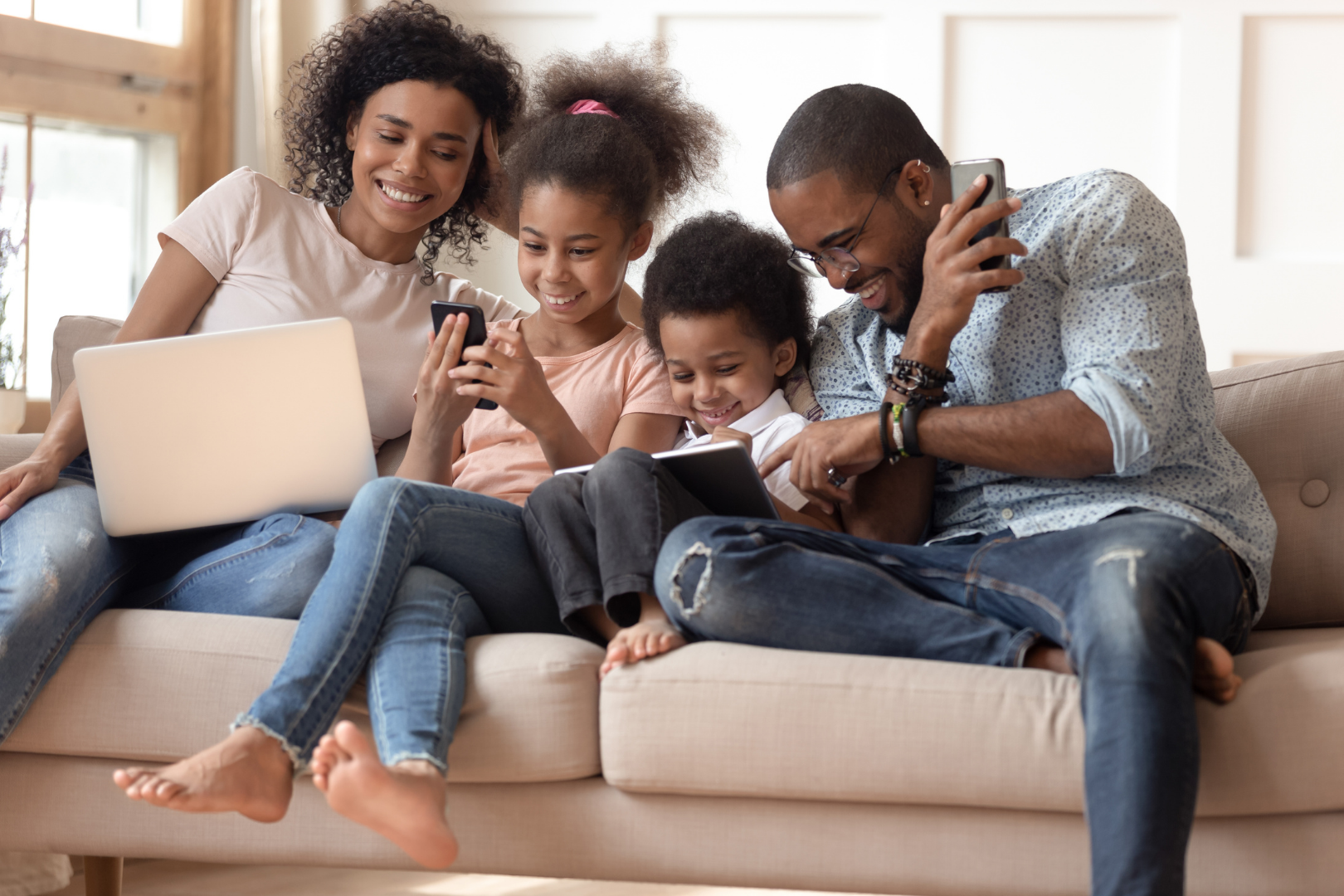 Family of four sitting on a couch, smiling and using a laptop and smartphones