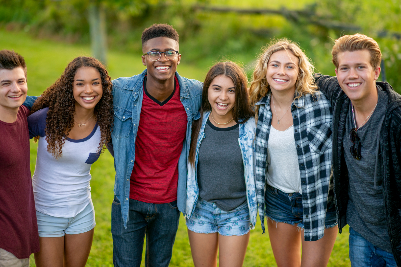 Five friends standing arm in arm outdoors on a sunny day, smiling at the camera.