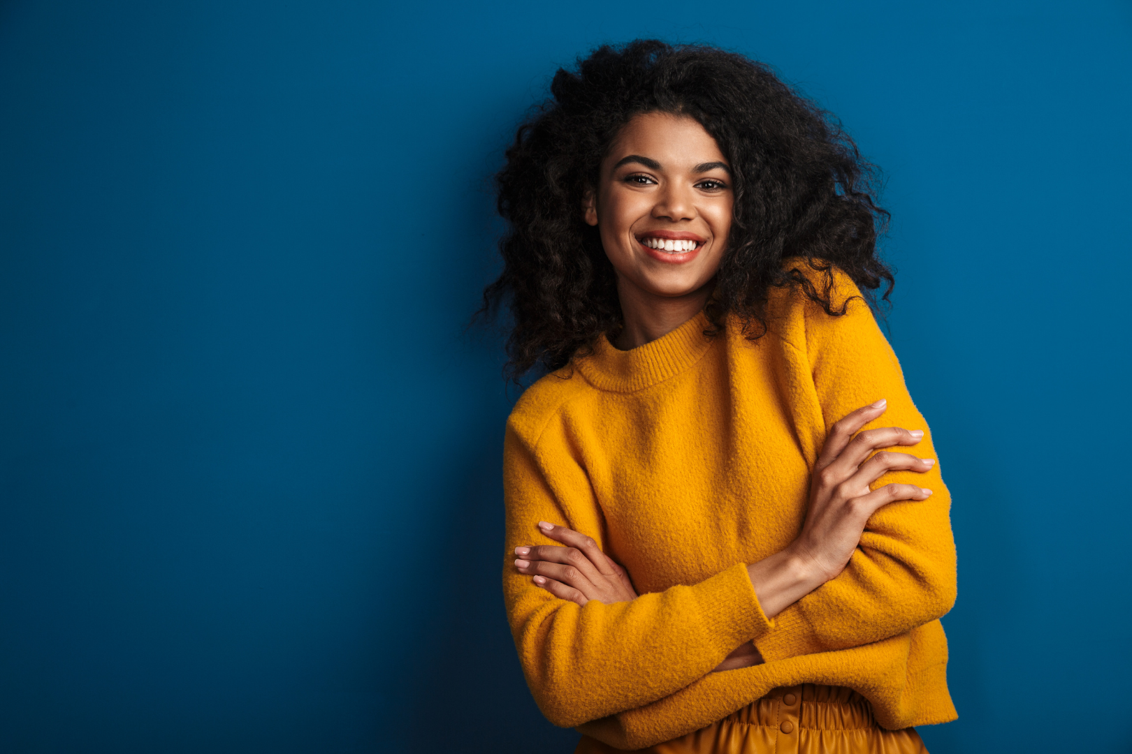 Smiling person with curly hair in a mustard sweater, arms crossed against a blue background
