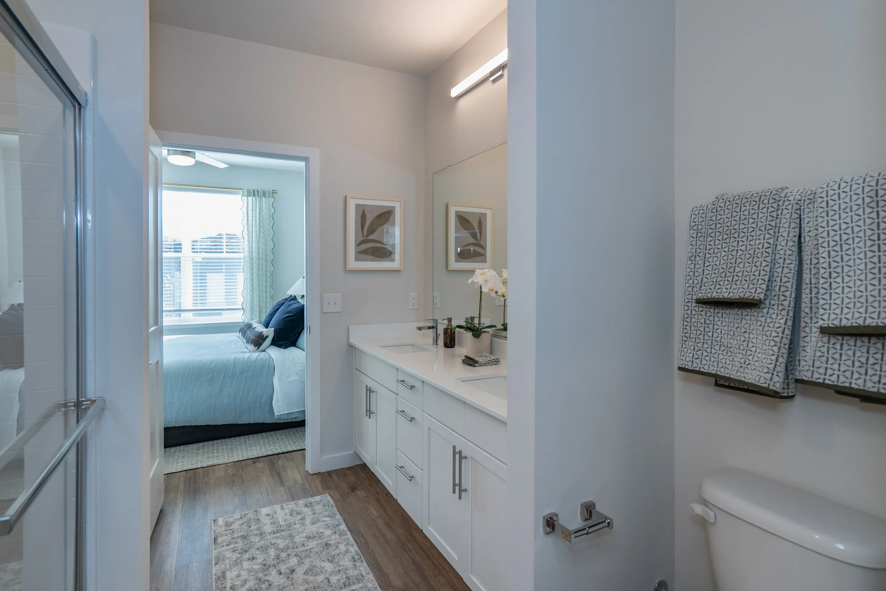 Modern bathroom with double vanity and glass-enclosed shower, opening to a bedroom with a large wind…