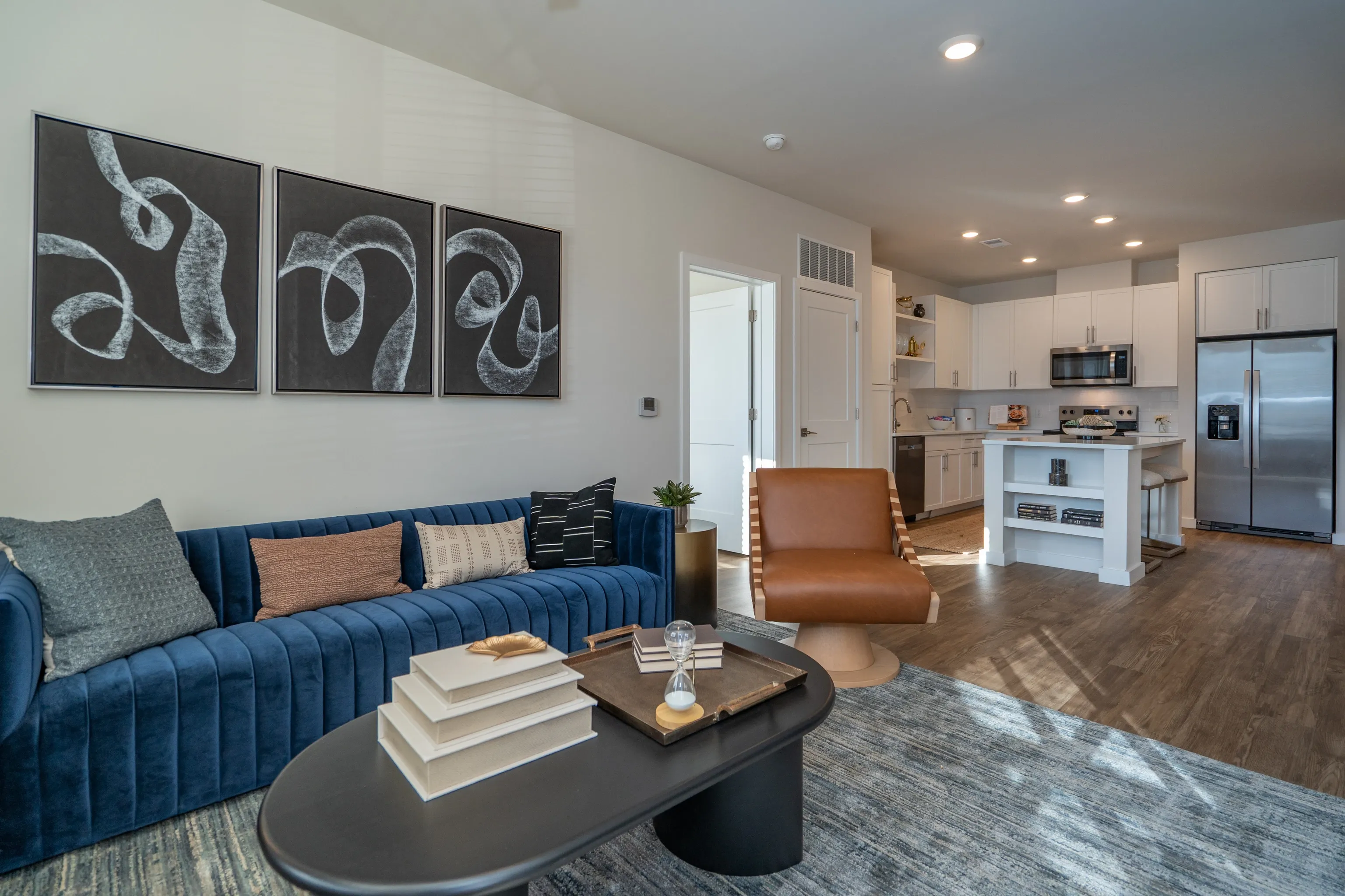 Modern living room with blue sofa, abstract art, and kitchen with island and stainless steel appliances.