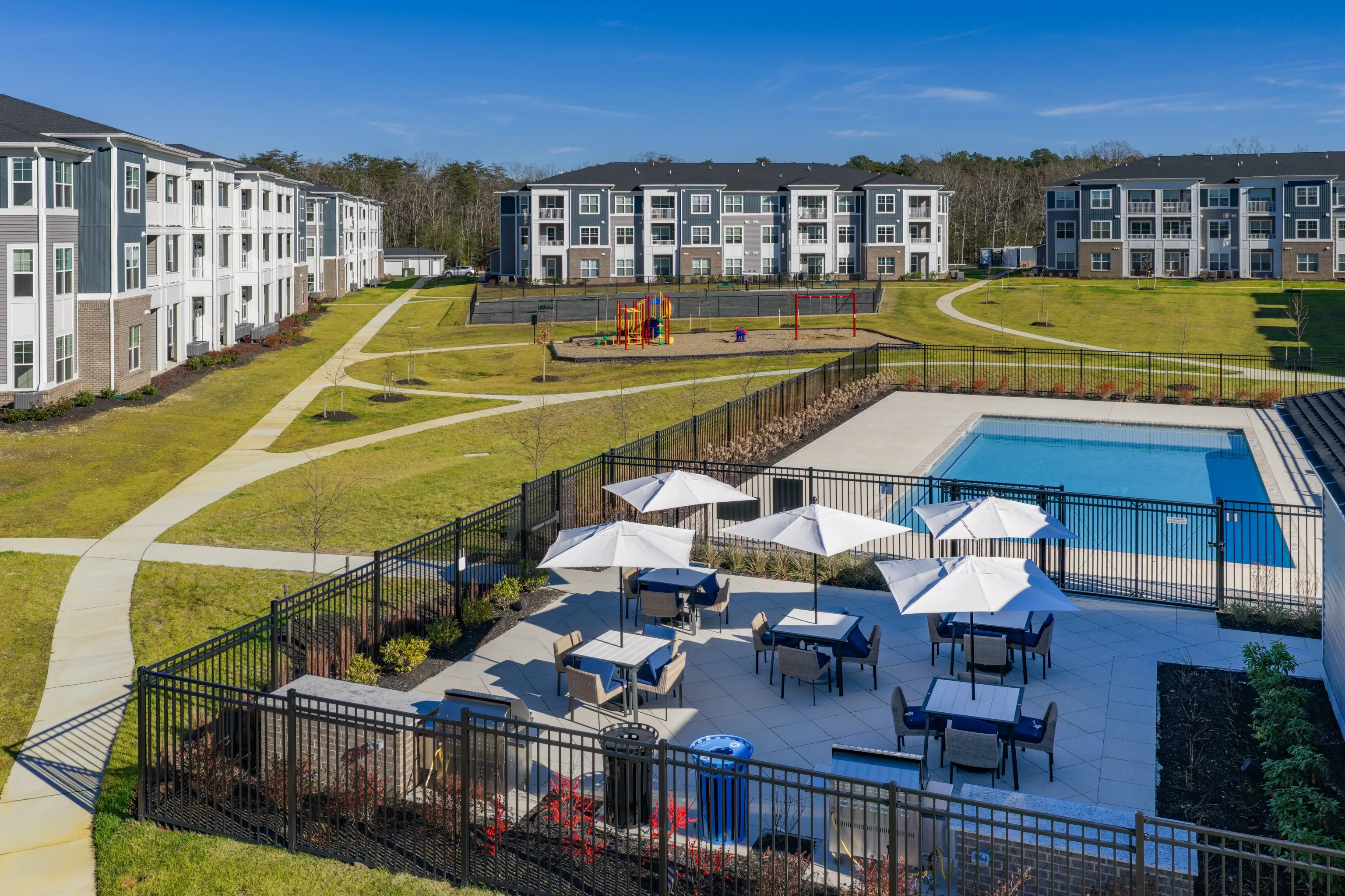 Outdoor swimming pool with patio seating and grills, adjacent to apartment buildings and playground.