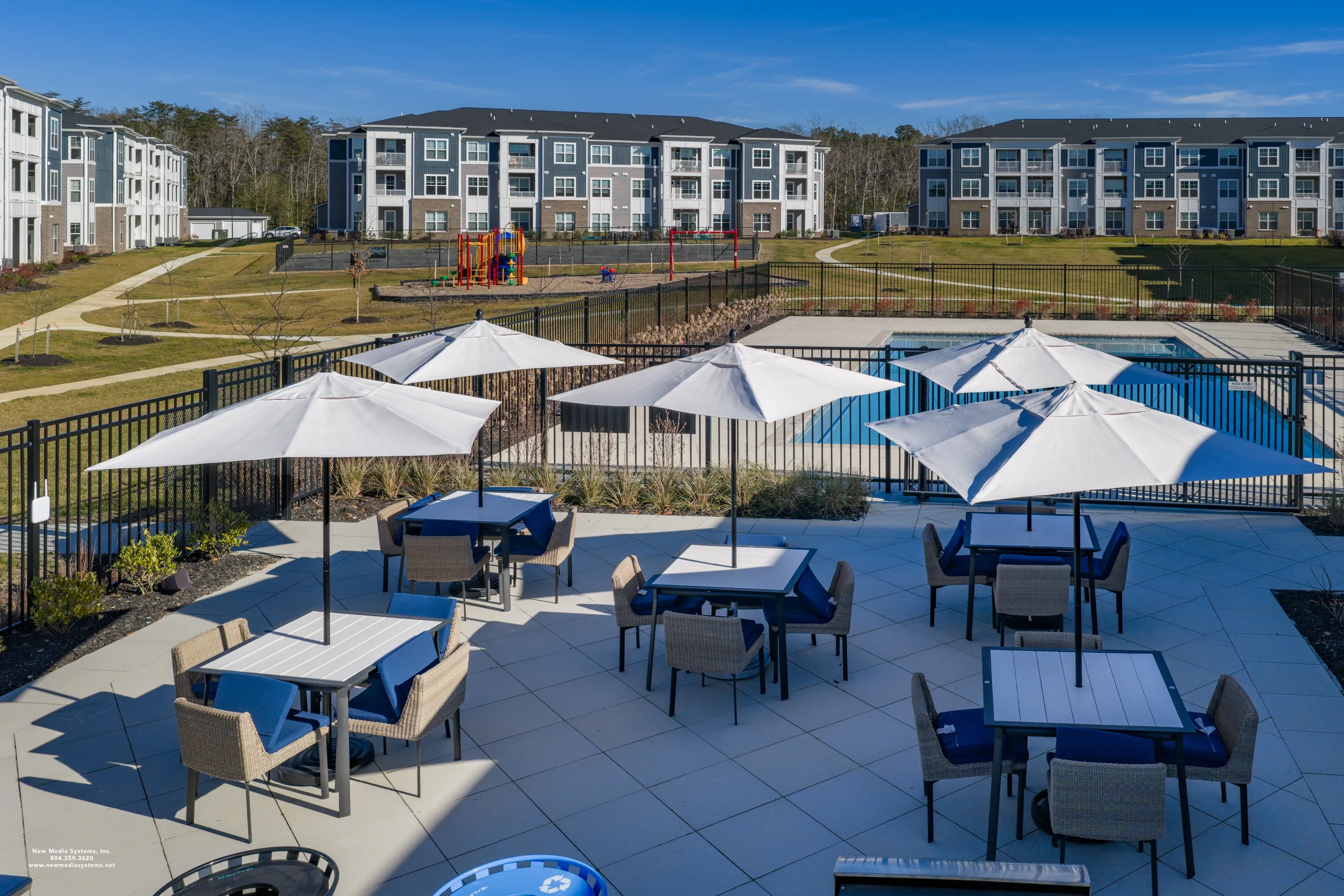 Outdoor seating area with tables and umbrellas next to a swimming pool and apartment buildings.