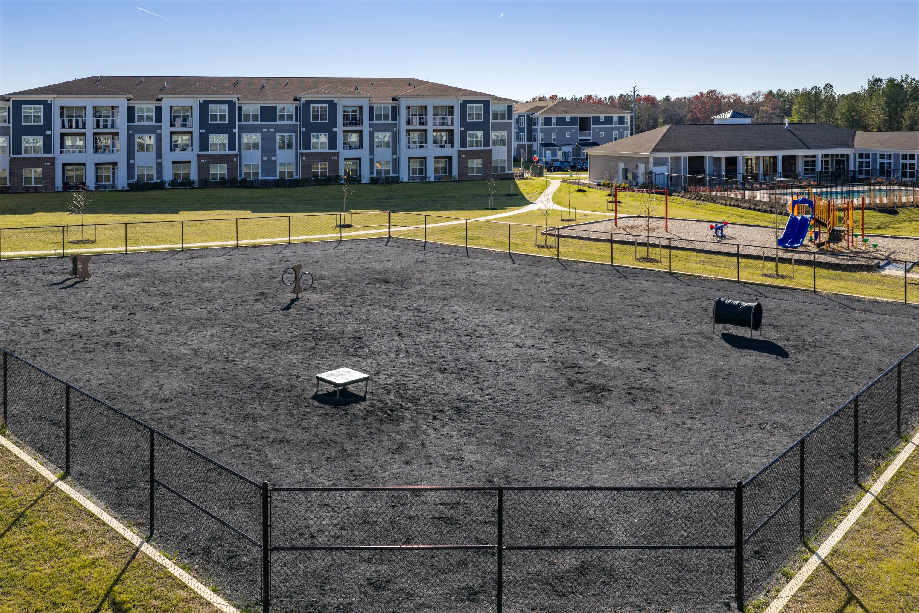 Fenced-in dog park with agility equipment and a playground in the background.