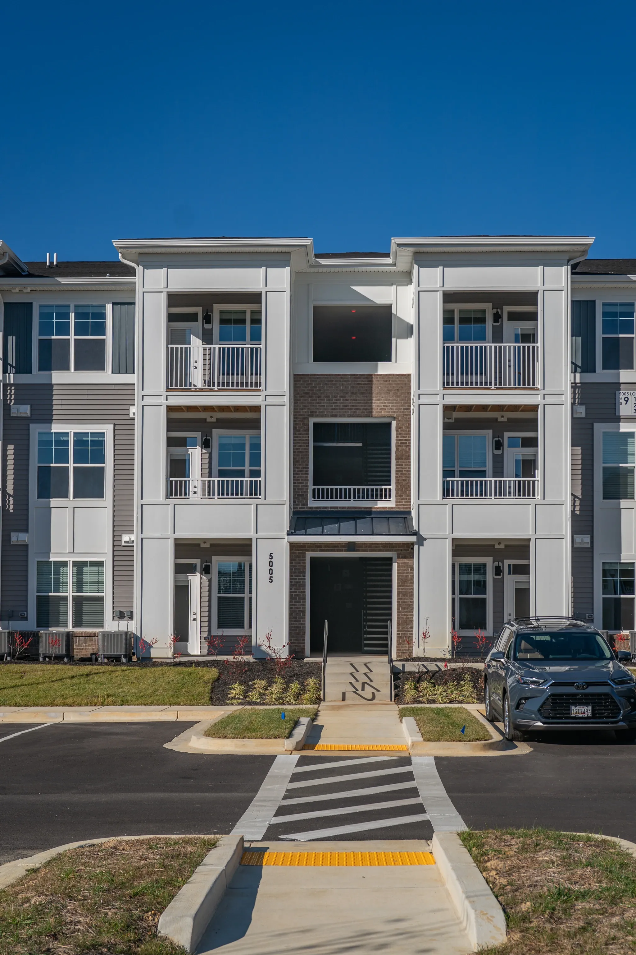 Modern apartment building exterior with balconies, windows, and a parking lot with a car.