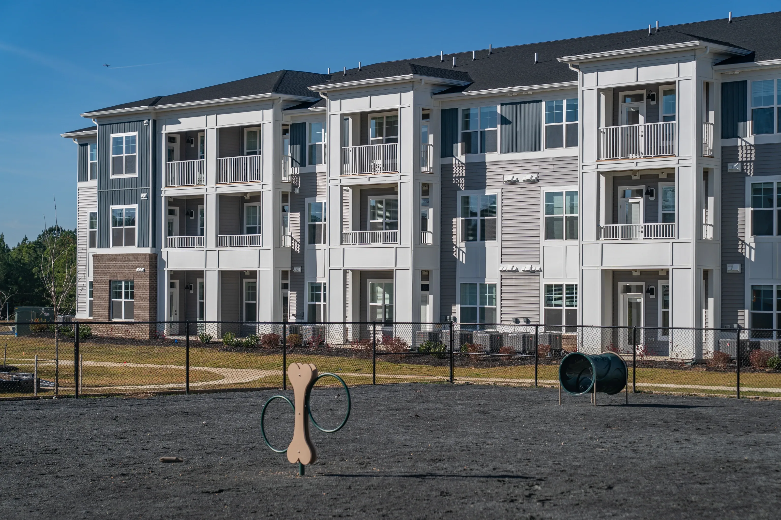 Exterior of a modern apartment building with balconies and a fenced dog park in the foreground.