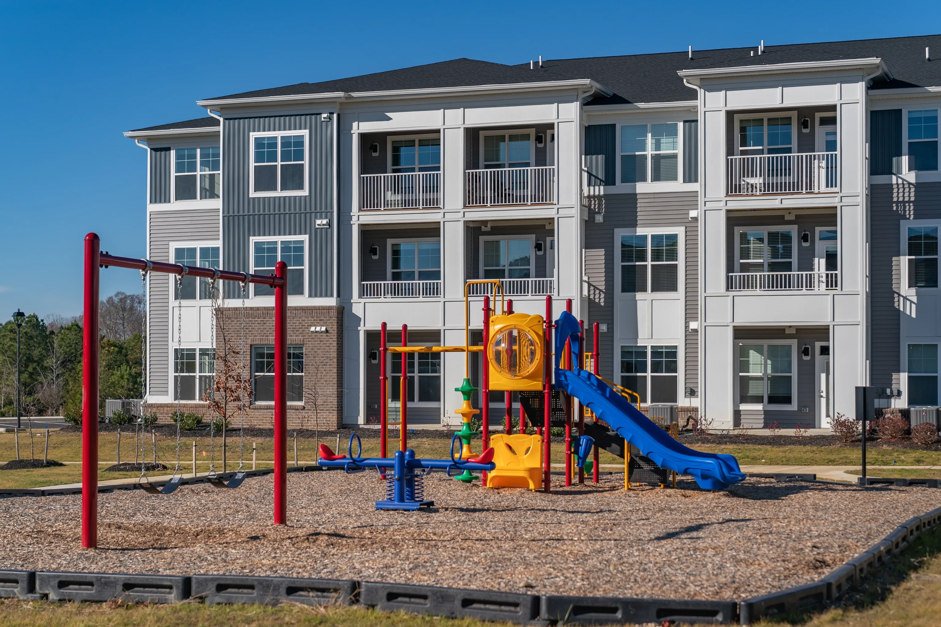 Playground with swings, slide, and seesaw in front of a modern apartment building.