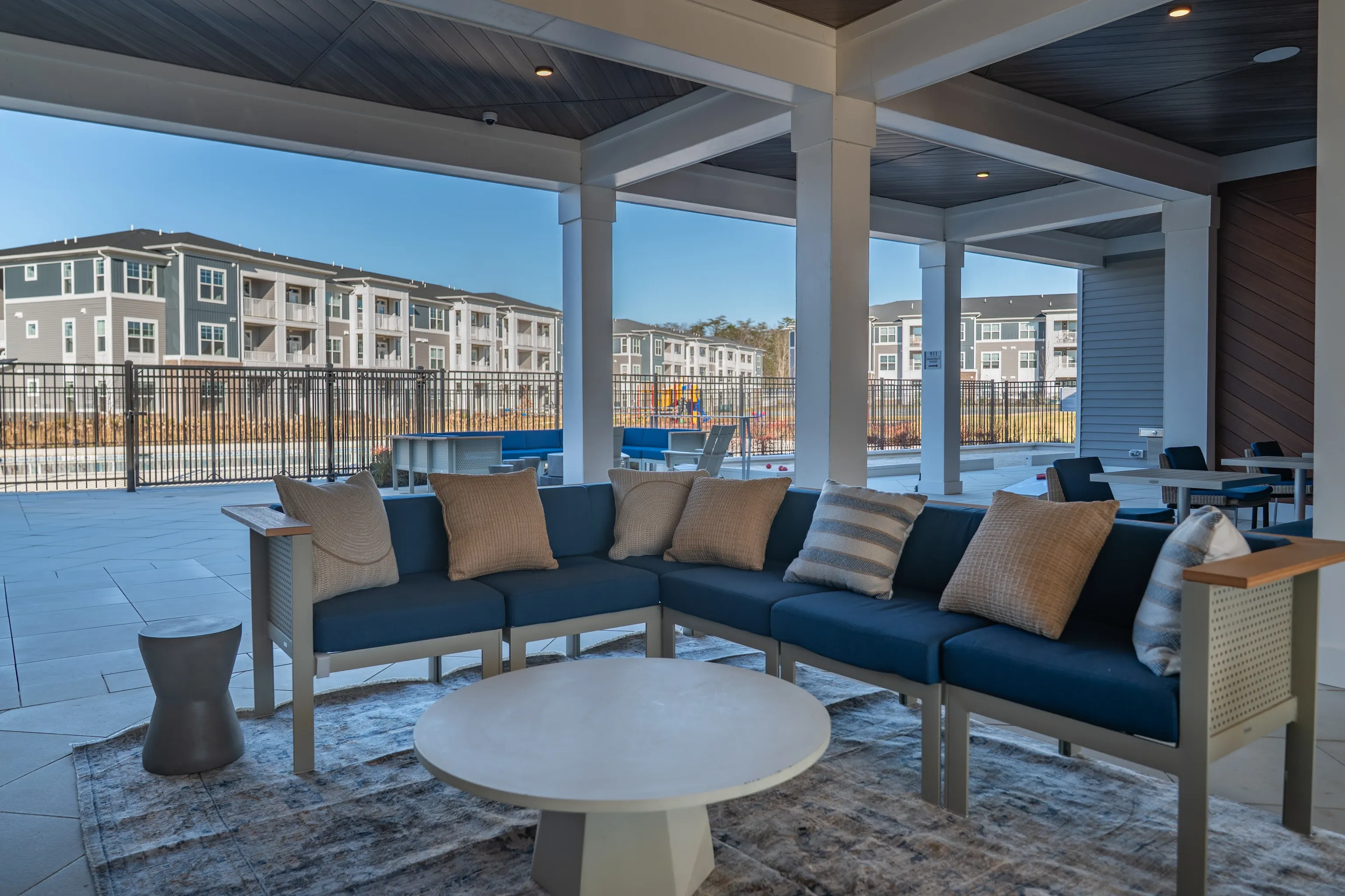 Covered outdoor seating area with blue sectional sofa and coffee table, overlooking apartment buildi…