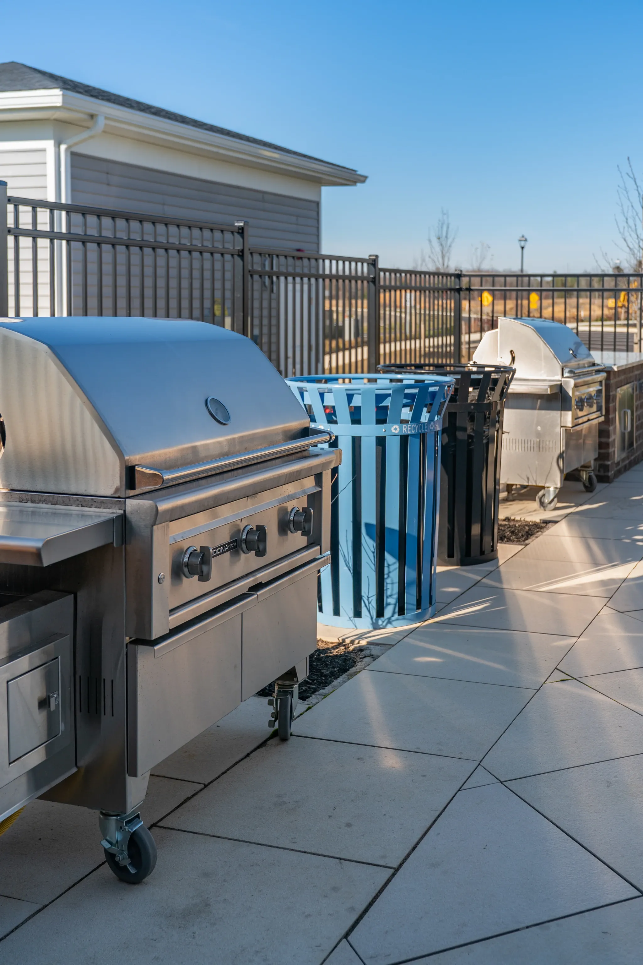 Outdoor grills and trash cans on a patio with a fence and building in the background.