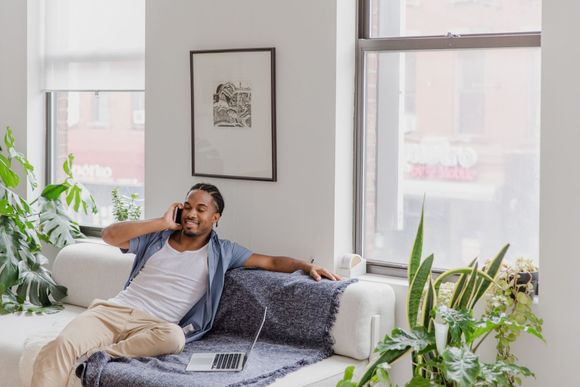 Person relaxing on a white sofa with a laptop and phone in a bright living room with plants.