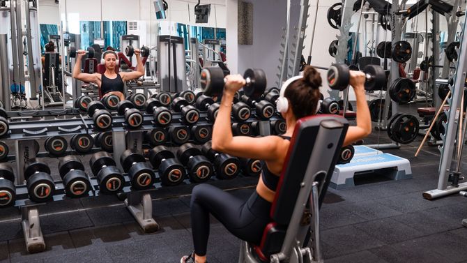 Person using a seated shoulder press machine in a gym, with dumbbells and mirrors in the background.