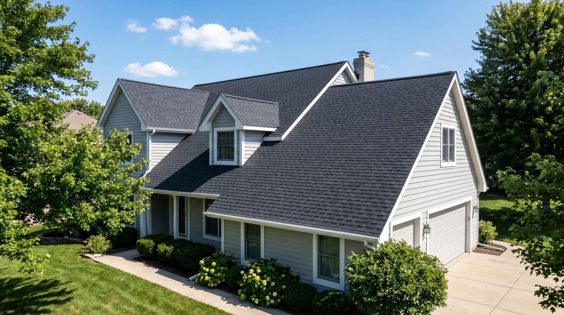 White suburban house with dark roof, dormer windows, and green trees on a sunny day