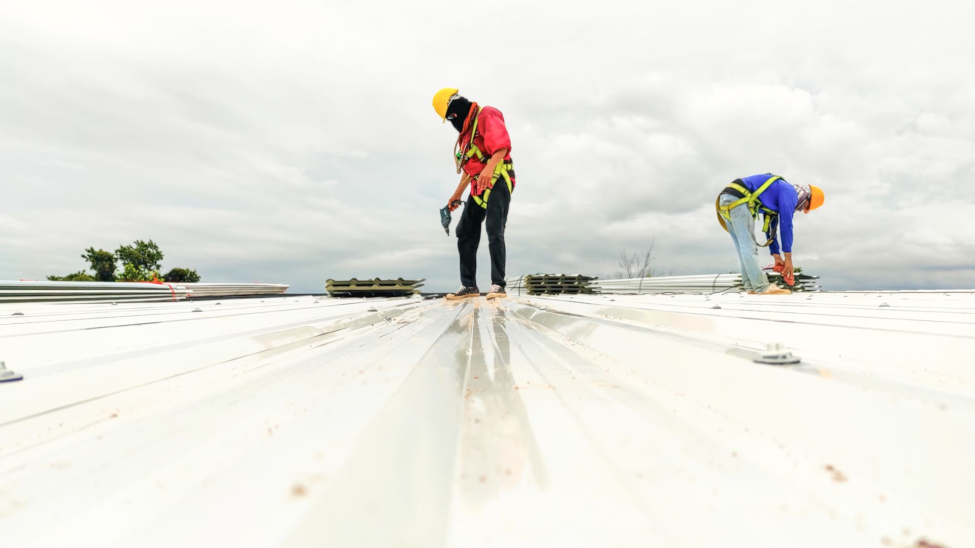 Two workers in safety harnesses work on a metal roof under cloudy skies.